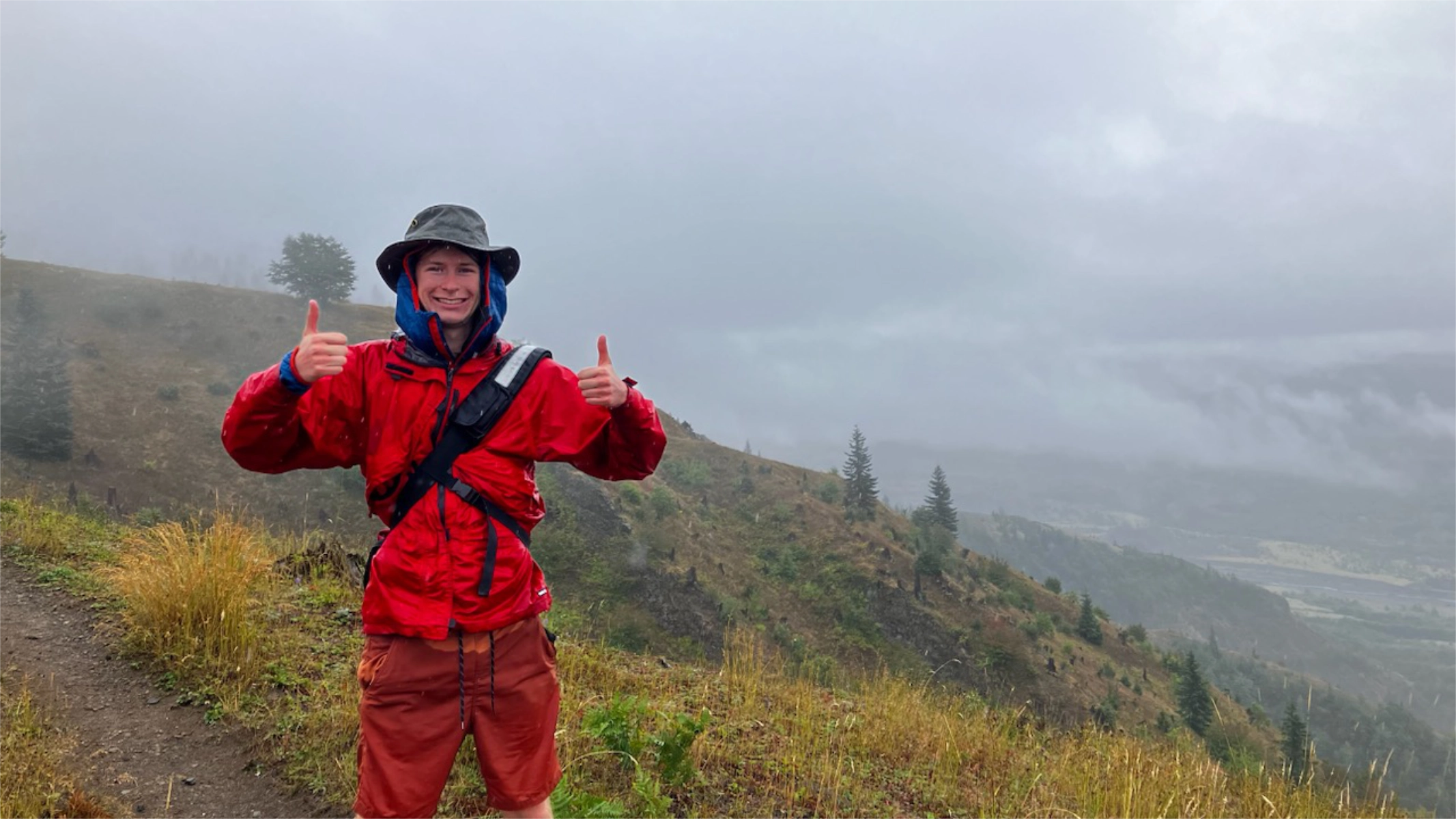 Photo of a person in a read rainjacket and a olive hat standing in the rain giving a double thumbs up.